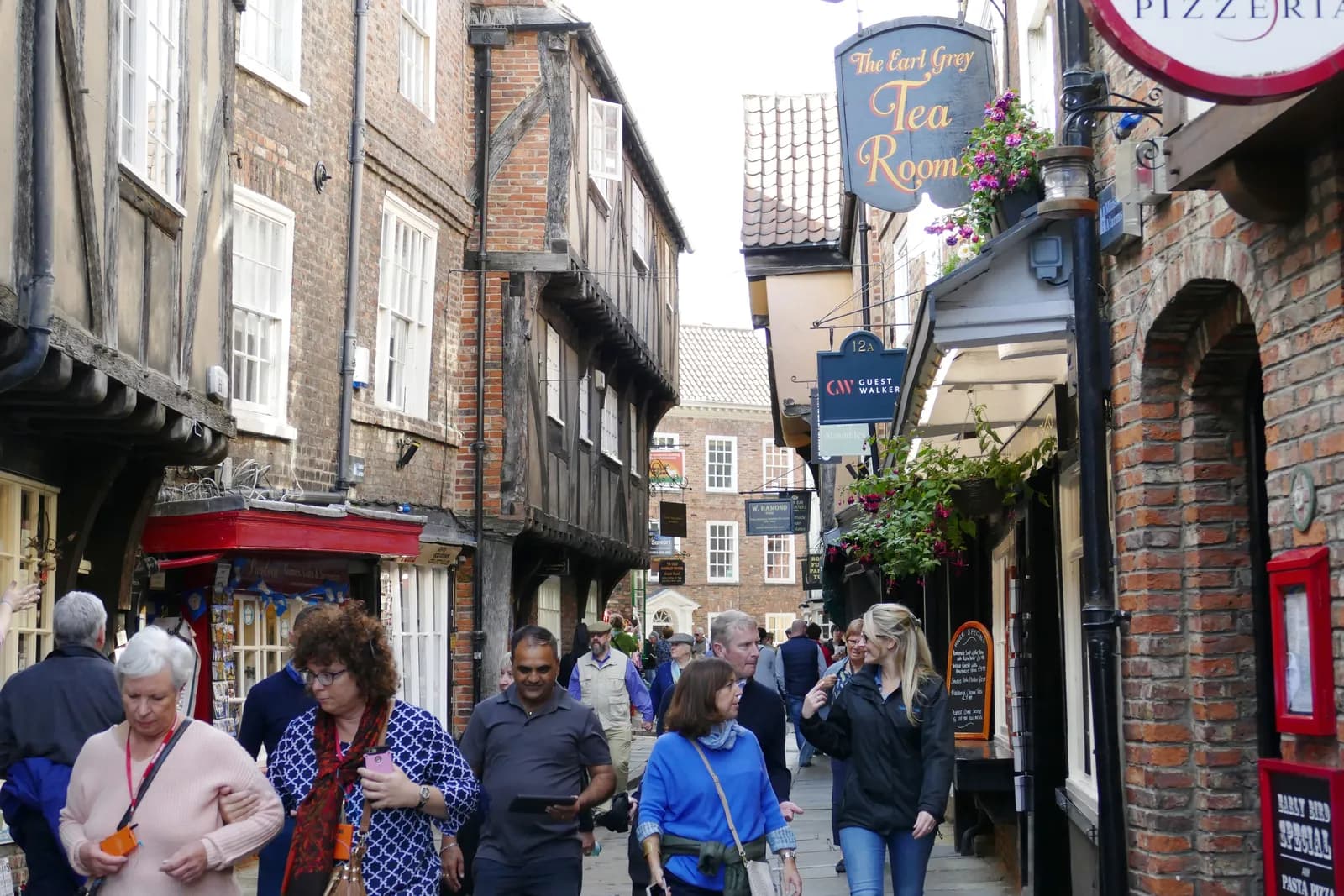 A bustling street in a historic town with people walking past old brick buildings with timber frames and shops, including 'The Earl Grey Tea Rooms'.