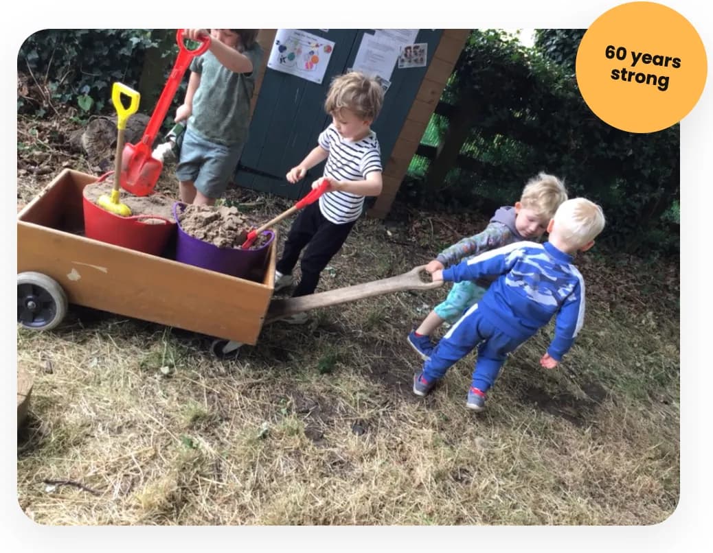 Four young children playing outdoors with a wooden wagon filled with sand, using shovels and pulling the wagon.
